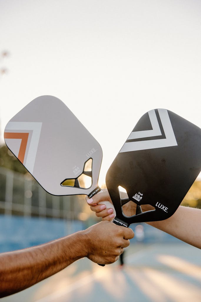 Two hands holding pickleball paddles outdoors during sunset.